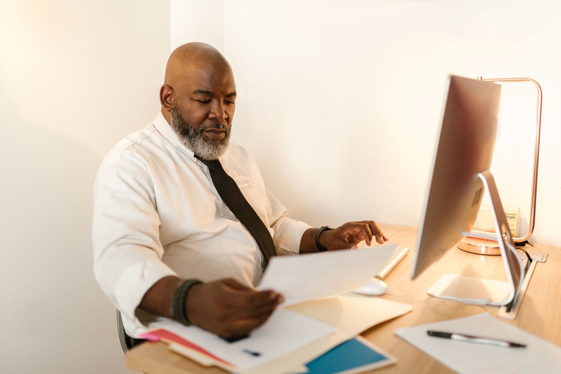 Bald man with beard in white shirt reviewing documents at desk with computer.